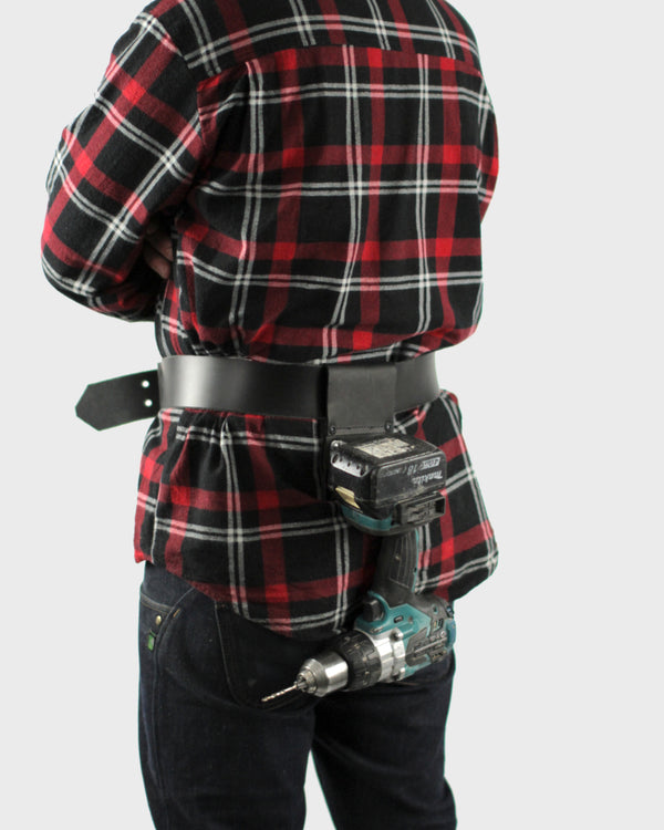 A person wearing a plaid shirt has a drill attached to a belt clip around their waist.